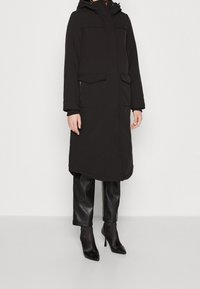 Manteau long noir avec capuche, deux poches avant et une poche zippée. Confectionné dans un tissu lisse et mat, assorti à un pantalon noir brillant et des talons.