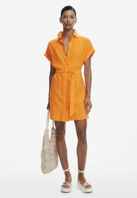 Abito arancione a maniche corte con vita cintrata, caratterizzato da dettagli ricamati. Abbinato a una borsa in crochet leggera e sandali platform spessi.