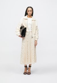 Crèmekleurig bomberjack met een gestructureerde kraag, wit overhemd met knopen, beige geplooide rok met zwarte stippen, zwarte handtas en zwarte sandalen met bandjes.