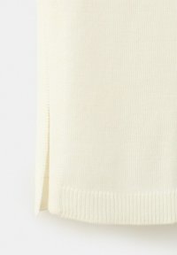 Približni prikaz donjeg ruba kremasto obojanog pletenog odjevnog predmeta s malim bočnim prorezom i rebrastim rubom.