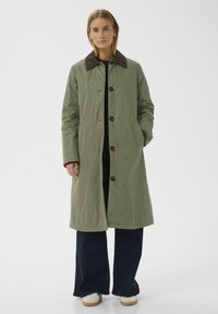 Olijfgroen lange jas met een bruine corduroy kraag, knopen aan de voorkant, zijzakken en een relaxte pasvorm, gecombineerd met donkerblauwe flared jeans.