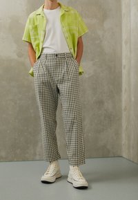 Person standing with hands in pockets wearing cream high-top sneakers, black-and-white checkered pants, white t-shirt, and lime green plaid short-sleeve shirt.