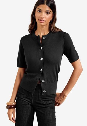 Junge Frau trägt eine eng anliegende schwarze Kurzarm-Strickjacke mit dekorativen Knöpfen und dunkle Jeans, posiert mit einer Hand in der Hüfte.