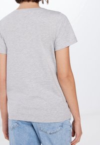 Persona vestida con una camiseta de manga corta gris claro y jeans de mezclilla azul claro, vista desde atrás contra un fondo blanco.