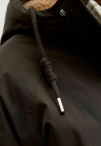 Brūna kapucīne jaka ar mīkstu, brūngani dzeltenu oderi. Tā ir aprīkota ar tumšu pinumu auklu ar metāla galu un spiedpogu aizdari ap kaklu.