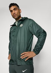Green, lightweight, windbreaker jacket with zipper closure, elastic cuffs, and hood. Features a circular logo and a Nike swoosh on the chest.