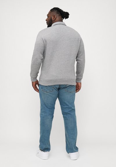 Szürke pulóver gallérral, hosszú ujjakkal és bordás mandzsettákkal, világoskék farmerral és fehér cipővel párosítva. Egyszerű, laza dizájn.