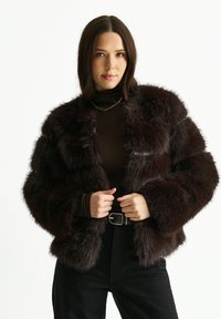 Brown faux fur jacket with a boxy design, plush texture, and wide sleeves. Styled with a dark turtleneck and high-waisted black pants.