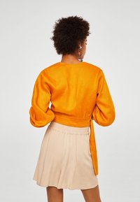 Femme aux cheveux bouclés portant un haut orange à manches longues et une jupe beige, vue de dos sur un fond uni.