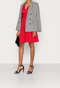 Robe rouge à col en V, taille cintrée et jupe évasée, associée à un blazer croisé en pied-de-poule noir et blanc et des talons noirs.