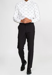 Camicia bianca a maniche lunghe con piccoli motivi di alberi verdi, abbinata a pantaloni neri slim e scarpe eleganti nere, con le mani intrecciate davanti.