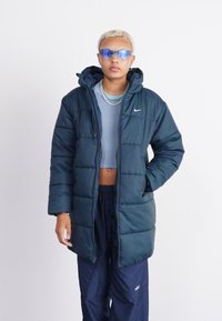 Κιτρινόμαυρο γεμάτο μπουφάν με κουκούλα, διαθέτει μπροστινό φερμουάρ και λογότυπο Nike. Συνδυάζεται με ένα κοντό τοπ και ναυτικές παντελόνες, φορώντας μπλε γυαλιά ηλίου.