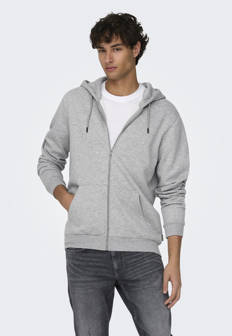 Grå zip-hoodie lavet af en bomuldsblanding, med en hætte som kan justeres med snore, frontlommer og ribbede manchetter. Båret over en hvid T-shirt.