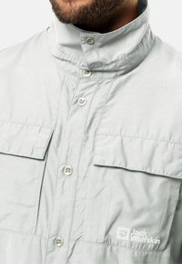 Camicia grigia chiara con colletto alto, due tasche sul petto e il logo "Jack Wolfskin" sul lato sinistro. Tessuto morbido e liscio.