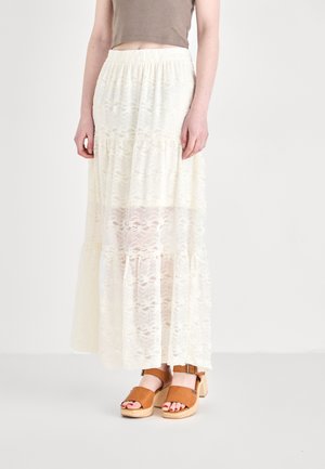 Jupe maxi blanche en dentelle avec un design à étages, texture transparente et taille élastique, associée à des sandales à plateforme marron.
