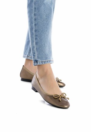 Bailarinas de bronce con punta redondeada, parte superior de tela, detalle de pequeño lazo y tacón bajo negro, combinadas con jeans azul claro con vuelta.