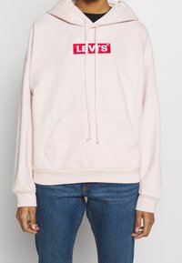 Světle růžová mikina s kapucí vyrobená z měkké látky, vybavená přední kapsou, šňůrkami a červeným nášivkou "LEVI'S" na hrudi.