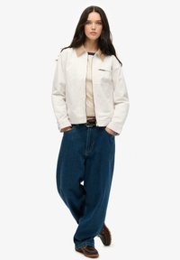 Hvid bomuldsjakke med tan krave, beige top og løse blå denimbukser. Brune sko med en tekstureret sål. Simpelt design.