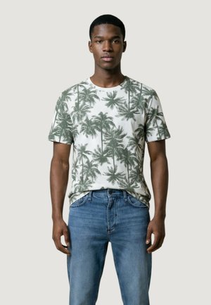 Giovane uomo che indossa una maglietta bianca con stampa di palme verdi e jeans blu, in piedi contro uno sfondo chiaro e uniforme.