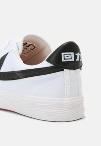 Hvit sneaker med et teksturert stoffoverdel, svart skinn hælflik med hvit skrift, og et mønstret gummisul.