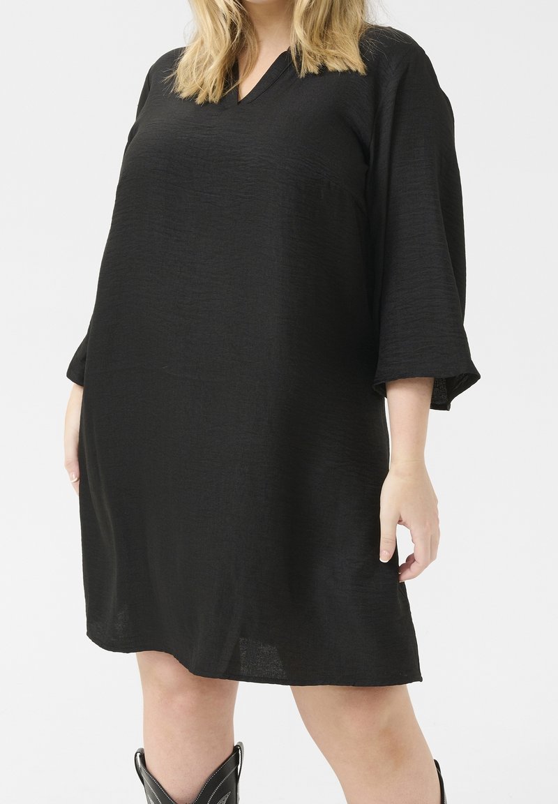 Femme portant une robe noire ample au genou avec des manches trois-quarts et des bottes noires mi-mollet, sur un fond clair uni.