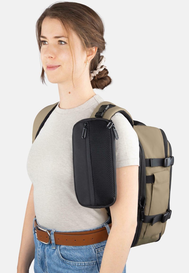 Rucksack mit beigem Hauptteil und schwarzen Akzenten, ausgestattet mit mehreren Reißverschlussfächern, verstellbaren Trägern und einer Netztasche an der Seite.