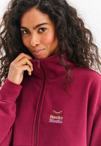 Bordowa bluza z kapturem na suwak, z wysokim kołnierzem, przednim zamkiem błyskawicznym oraz haftowanym logo „Naoko Studio” z motywem oka w kolorze złotym i wielobarwnymi akcentami.