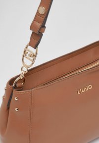 Sac à main en cuir marron à texture lisse, doté d'une fermeture éclair dorée, d'un logo, et de clous décoratifs dorés sur le côté. Bandoulière réglable incluse.