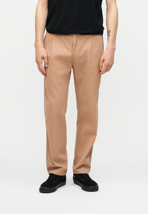 Homme portant un pantalon beige à jambes droites, des baskets noires et une chemise noire, debout avec les mains détendues le long du corps sur un fond uni.