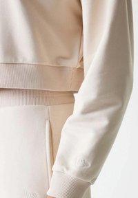 Felpa corta beige con orlo e polsini a coste, caratterizzata da un logo ricamato sulla manica. Tessuto morbido con una texture liscia.