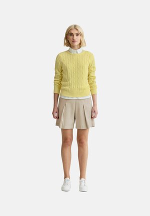 Kvinde stående iført en gul kabelstrikket sweater over en hvid skjorte med krave, beige plisserede shorts og hvide sneakers på en hvid baggrund.