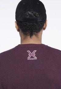 Felpa bordeaux con scollatura a girocollo, caratterizzata da un piccolo logo ricamato in rosa e bianco sulla parte posteriore vicino alla scollatura. Cappello in cord pubblico nero.