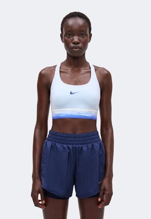 Atletische vrouw die een witte Nike sport-bh en blauwe Nike shorts draagt, staat tegen een eenvoudige lichte achtergrond.