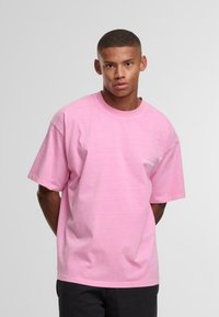 Noor mees lühikeste juustega, seljas lõdva heleda roosa T-särk ja mustad püksid, seismas käed selja taga tagasi, valge taustaga.