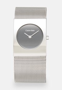 Montre avec bracelet en maille argentée, présentant un boîtier rectangulaire, un cadran gris et des aiguilles des heures et des minutes minimalistes blanches. Logo de la marque affiché à 12 heures.
