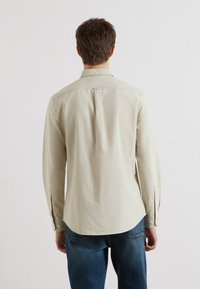 Beige langslopen shirt met een puntkraag, rugplooi en knoopmanchetten. Soepele textuur met een ronde zoom en een getailleerde pasvorm.