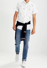 Kurzärmeliges weißes Hemd mit grünem Palmenmuster, kombiniert mit blauen zerrissenen Jeans und schwarzem Pullover, der in der Taille gebunden ist; weiße Sneakers.