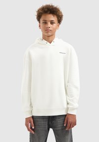 Tienerjongen met krullend haar, gekleed in een witte hoodie met kleine "BALLIN" tekst en vervaagde zwarte jeans, tegen een effen grijze achtergrond.