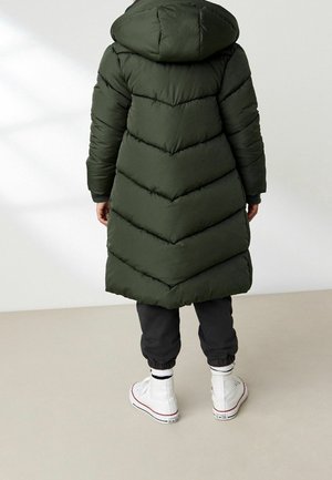Manteau matelassé vert foncé avec capuche, présentant un design en chevrons, associé à un pantalon noir et des baskets montantes blanches.