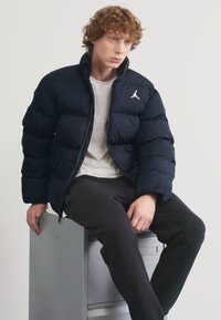 Mornarska puhovka z zadrgo in izvezenim logotipom Jordan. Model nosi belo srajco in črne hlače, sedi na sivi omari za dokumente.