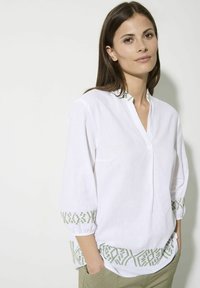 Weißes Leinenbluse mit grünen gestickten Akzenten am Kragen und an den Bündchen, mit einem V-Ausschnitt und dreiviertellangen Ärmeln.