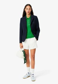 Marinblazer, grön polo, vita shorts och ljusblå sneakers. Vit handväska med en grön scarf som är knuten på handtaget.