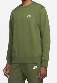 Man som bär en olivgrön Nike sweatshirt och matchande sweatpants med vita Nike-logotyper på bröstet och låret.