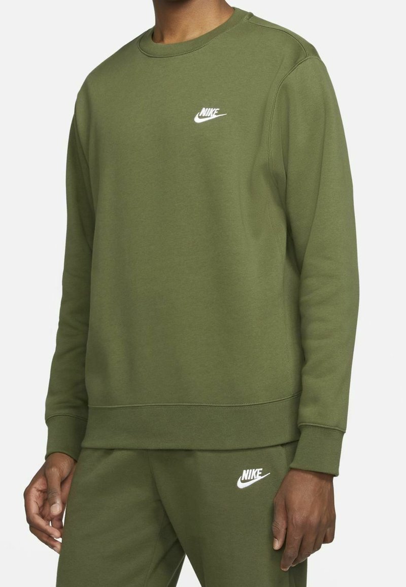 Man som bär en olivgrön Nike sweatshirt och matchande sweatpants med vita Nike-logotyper på bröstet och låret.