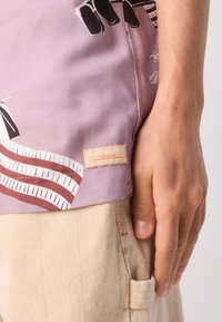 Kortermet, lavendel skjorte med abstrakte mønstre, med et stoffetikett som har "Scotch & Soda" logo, parret med beige bukser.
