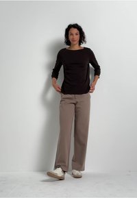 Top marrón de manga larga combinado con pantalones beiges de pierna ancha. Zapatillas blancas con acentos en marrón claro. Fondo liso.