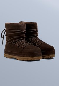 A barna szarvasbőr csizmák bokáig érő kialakítással, texturált anyagokkal, vastag gumitalppal, mintázott mintával és fűzőrészletekkel rendelkeznek.