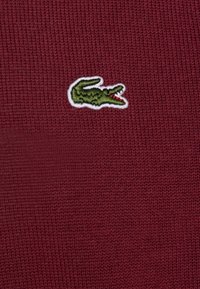 Tessuto a maglia color marrone scuro con un logo di coccodrillo verde bordato di bianco, che mette in evidenza la superficie testurizzata e i dettagli chiari del simbolo.