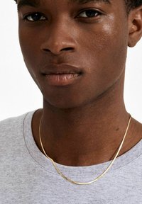 Guld kæde halskæde på en person iført en grå T-shirt. Halskæden har et glat, tyndt design; huden er mørk med en naturlig tekstur.