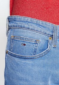 Jeans de mezclilla en azul claro con un acabado desgastado. Presentan un pequeño bolsillo trasero acentuado por un parche de logo en rojo, blanco y azul.
